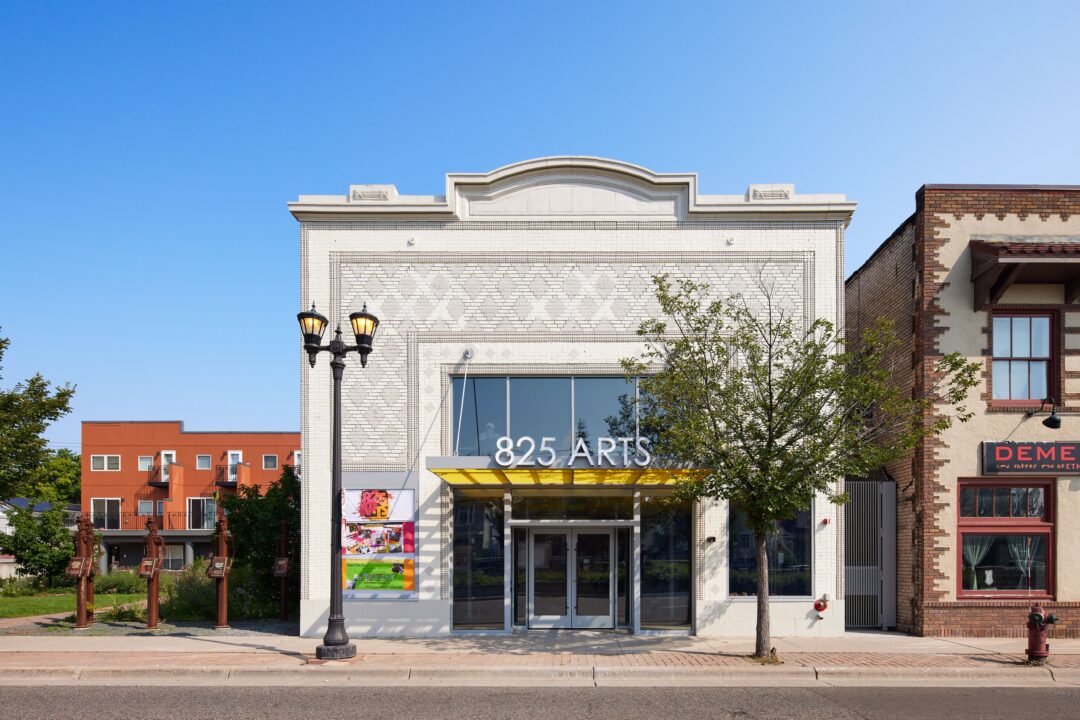 a front view of 825 Arts; the building is a 1915 Beaux Arts style theater with modern conversions, including large storefront windows, a yellow marquee, and a sign reading "825 Arts". In front of it are a sidewalk and University Avenue. To one side is a small green space.