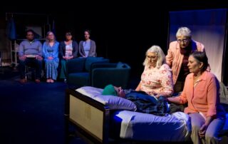 A group of people sit at the bedside of a person. An audience is seated in the background.