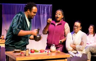 A person in a green track suit pours tea for a person in a pink zip up sweater. She is smiling, with her mouth open, as if mid-sentence.