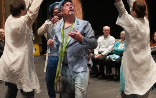 A group of robed spirits wave a scarf over a befuddled and amazed person, who walks through them.