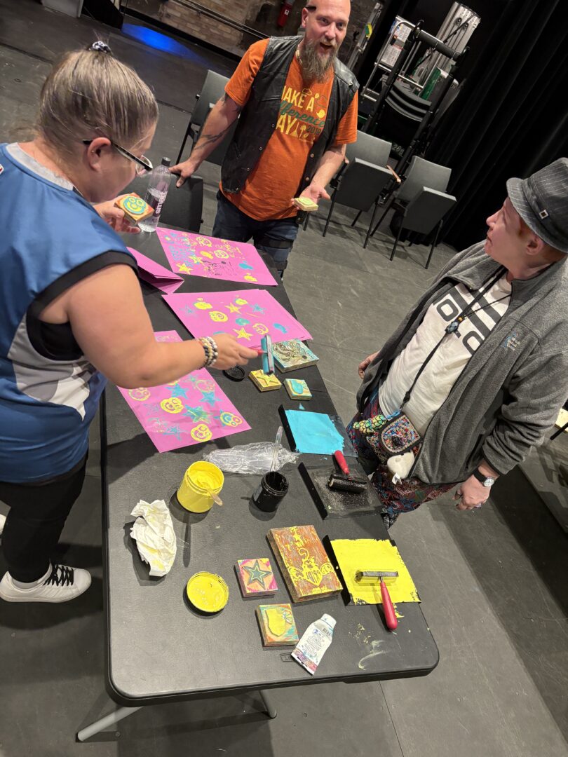 Three people stand around a folding table, on which sits block printing supplies and paper, which is printed with blue and yellow designs. 