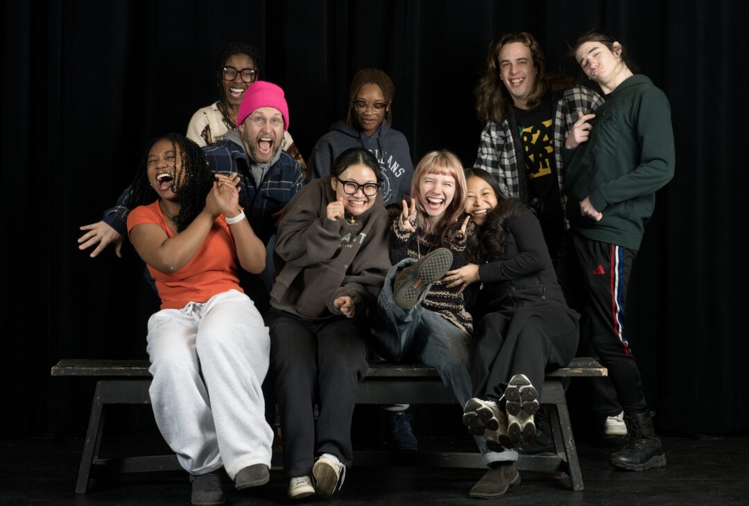 Family Portrait at 825 Arts An image of the Yalt team, running to sit down in the frame. Everyone is smiling, and Charity, on the far left, laughs with her hands extended in front of her. Lucy throws up to peace signs on the righ, sandwiched between Ku ku and Som La. Tyler is behind charity, arms spread and smile wide. Behind him is Mahogany, also smiling and laughing. to their right, in the center, is Myra, who looks down at those in front of her, smiling. Next to her, Ethan and Miles pose.