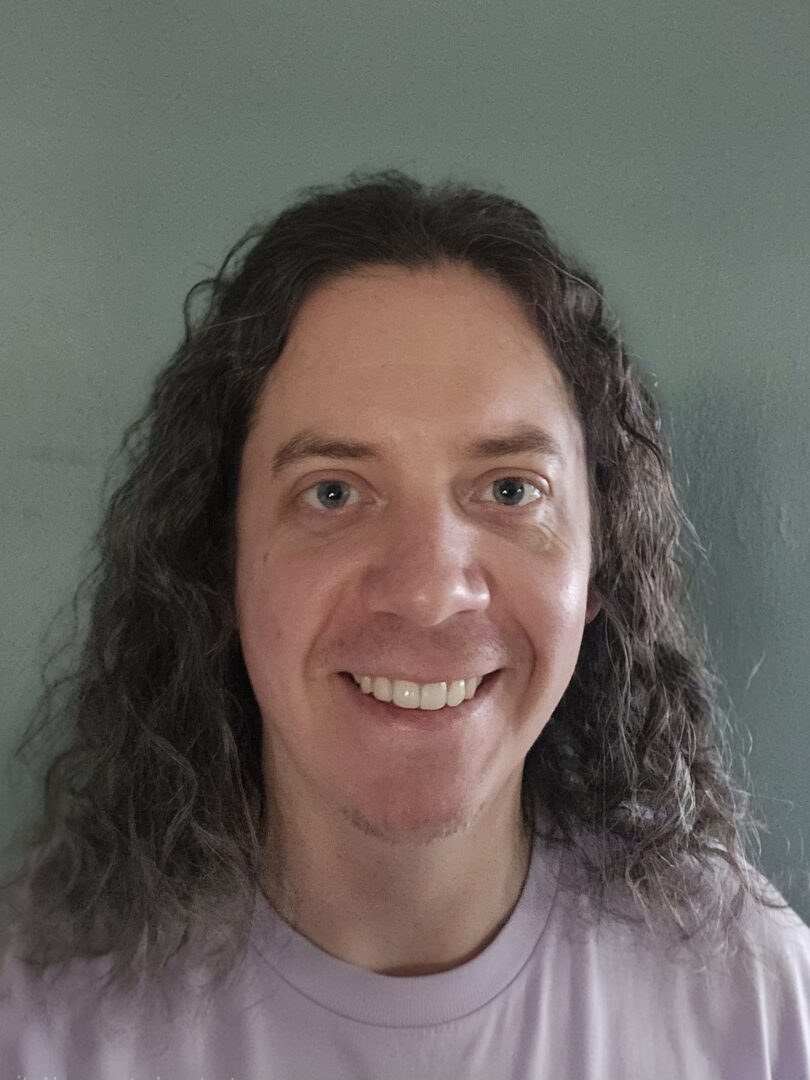 1000013937 Sabastian Boyle-Mejia, a pale-skinned, curly haired person in a pink shirt, who smiles at the camera.