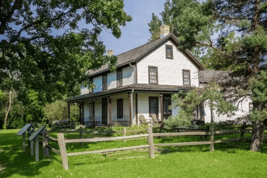 Gibbs farm, a white and grey three-story farmhouse, with a tamarack fence surrounded by trees.